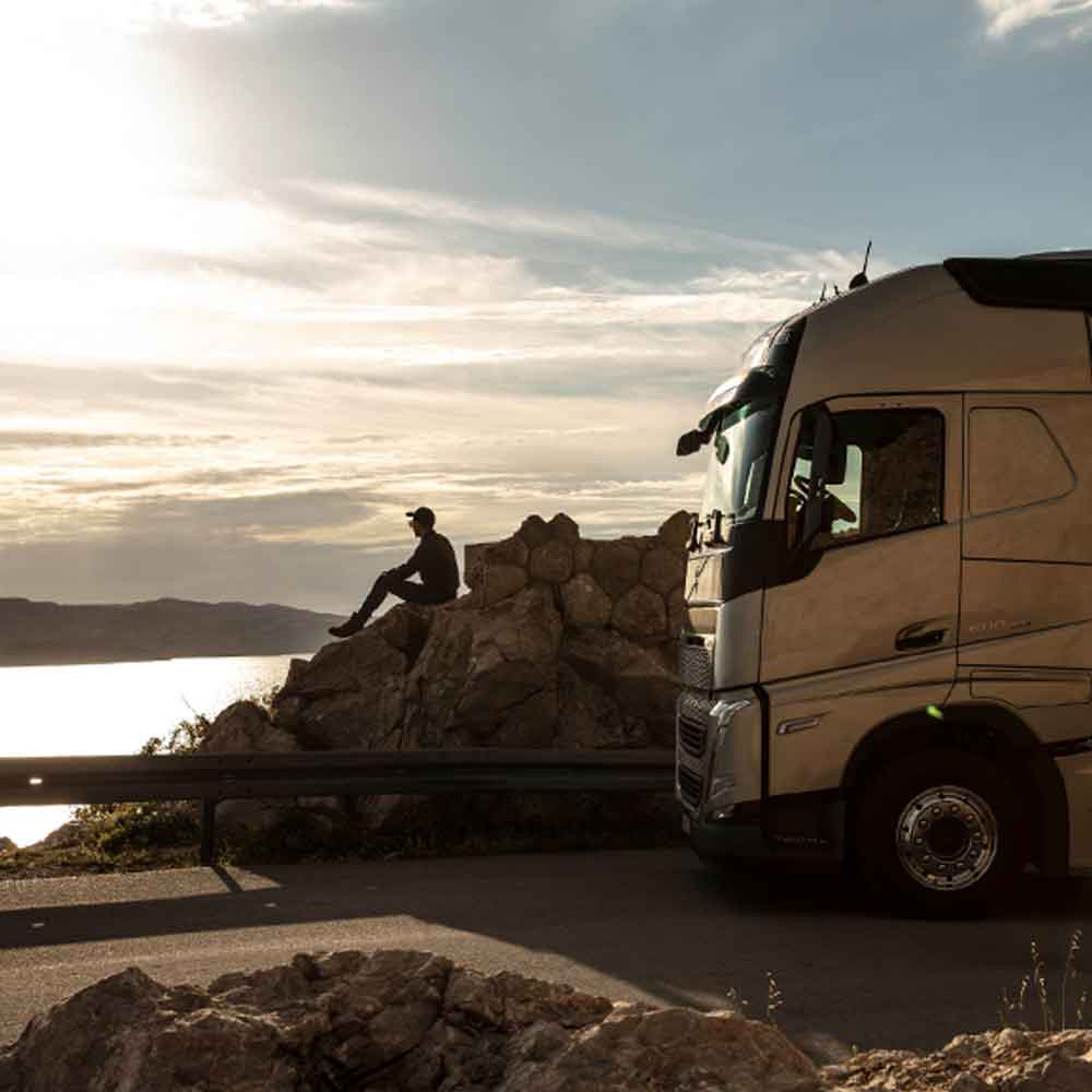 Ein Mann sitzt auf einem Felsen und sieht bei Sonnenuntergang auf das Meer hinaus, während sein Volvo FH neben ihm abgestellt ist